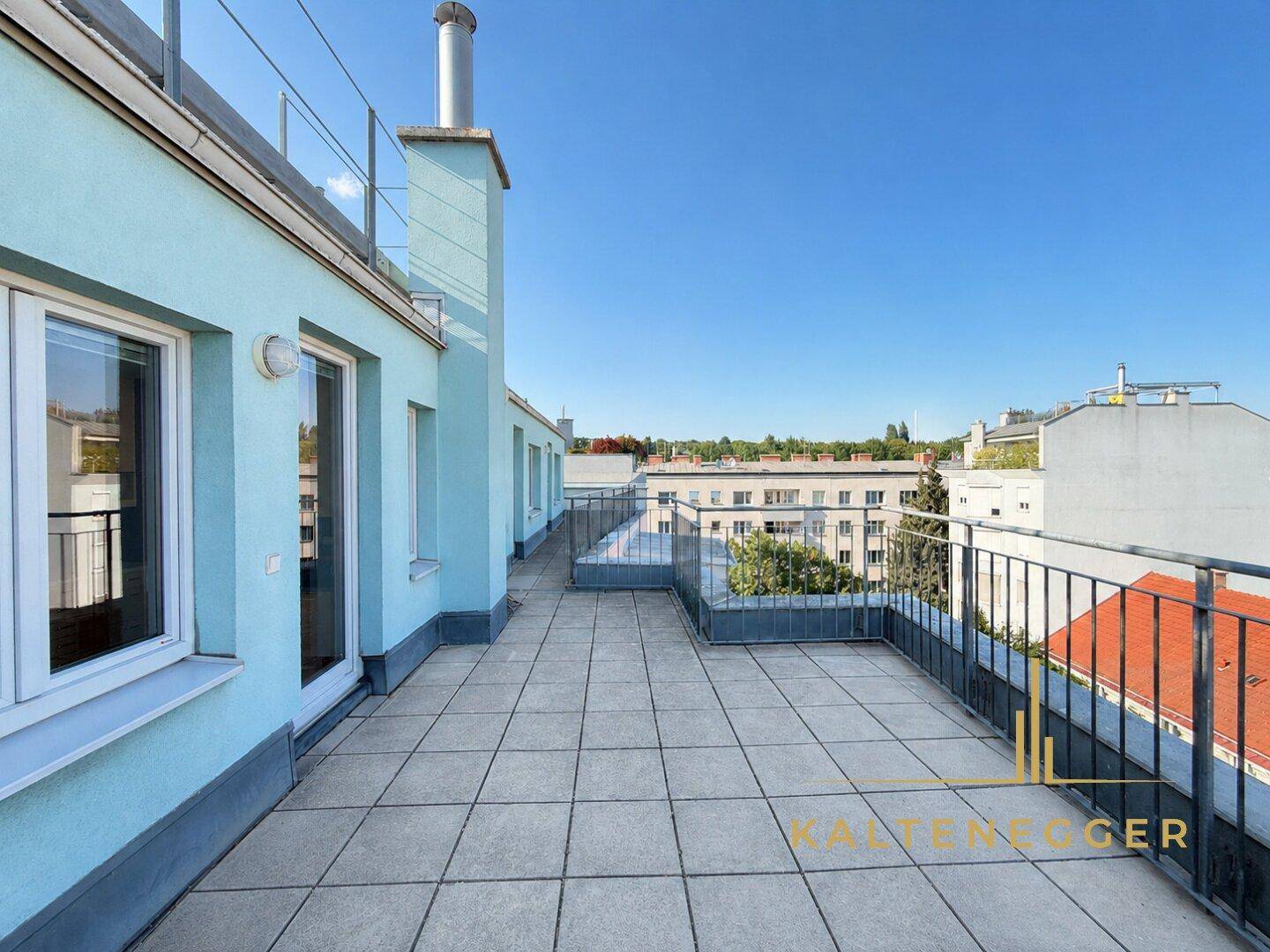 Lange Dachterrasse mit Fliesenboden und Metallgeländer, umgeben von Stadtansichten.