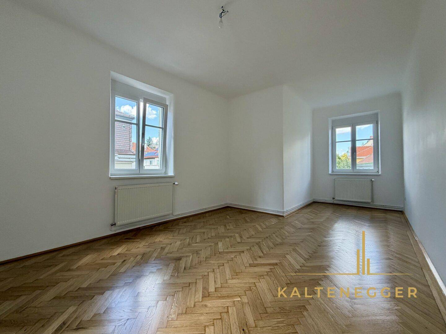 Helles Zimmer mit Fischgrätparkett und zwei Fenstern, die viel Tageslicht hereinlassen.