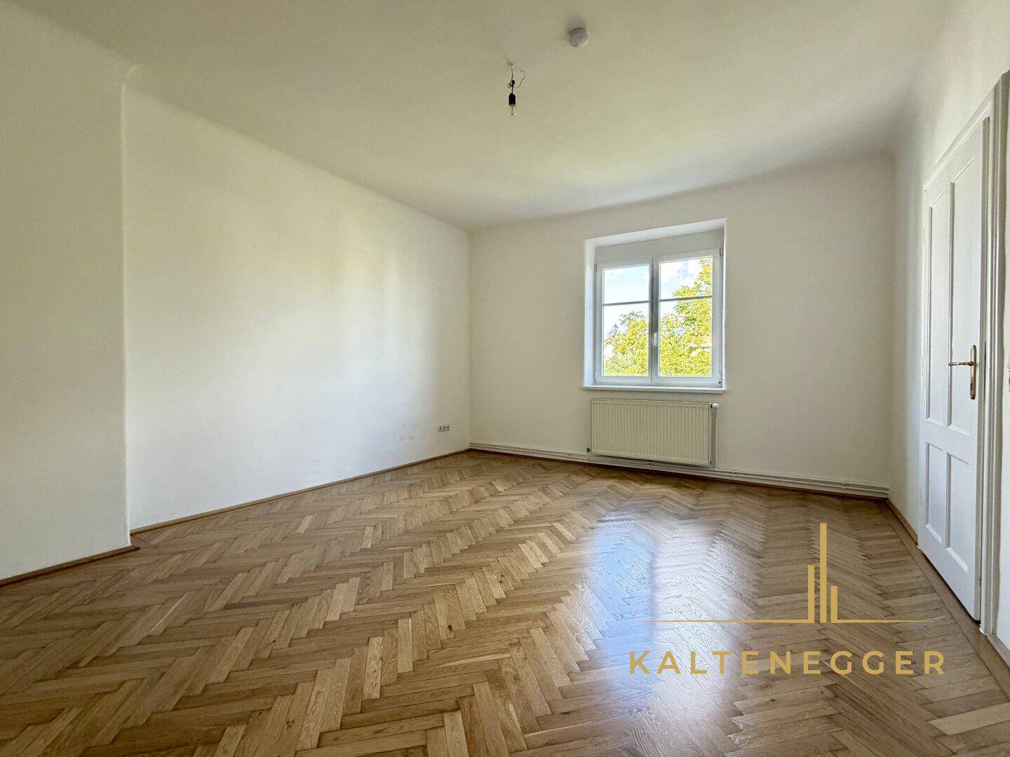 Geräumiges Zimmer mit klassischem Fischgrätparkett und einem großen Fenster mit Blick ins Grüne.