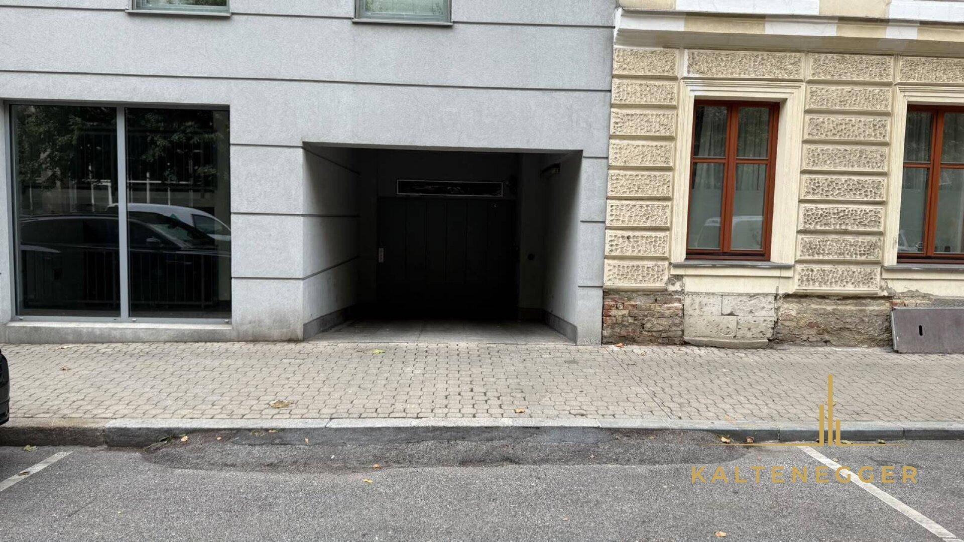 Eingang zur Tiefgarage an der Gebäudefassade mit Pflastersteinen und angrenzendem Altbau an der Straße.