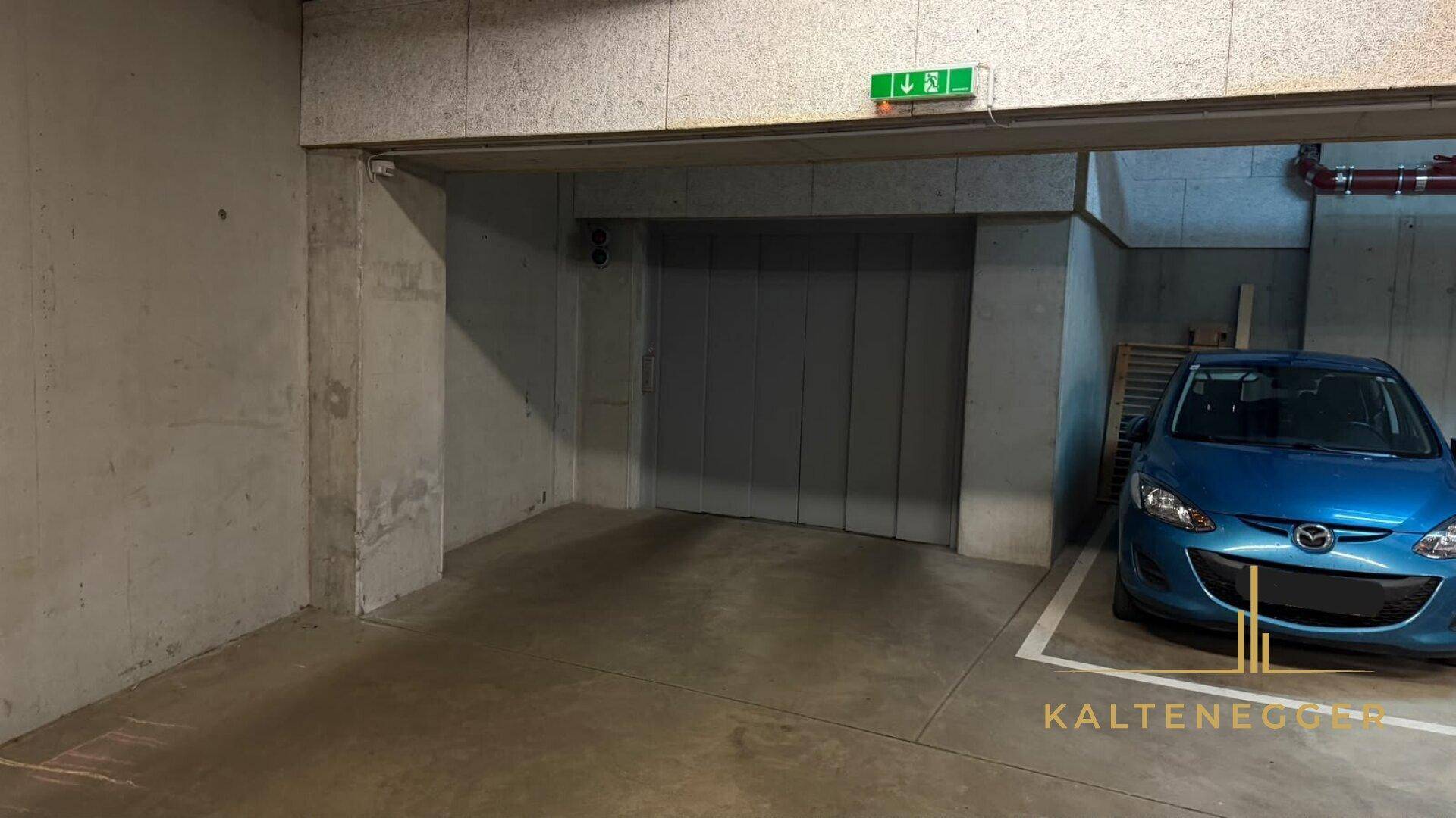 Tiefgarage mit einem blauen Auto, einem Aufzug und Betonwänden sowie sichtbaren roten Leitungen an der Decke.
