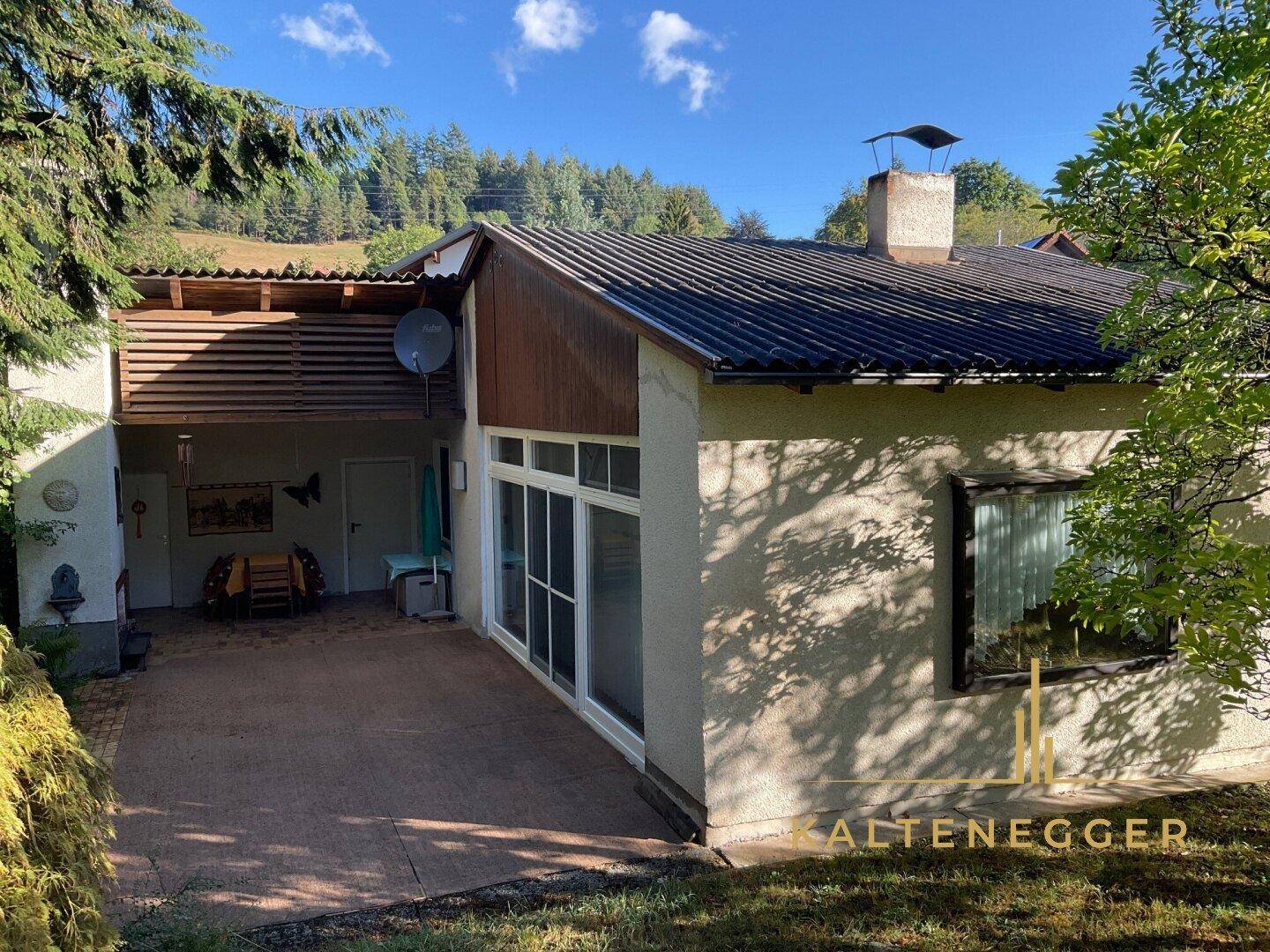 Rückseite des Hauses mit überdachtem Terrassenbereich, gepflastertem Boden und Blick auf die Hügel.