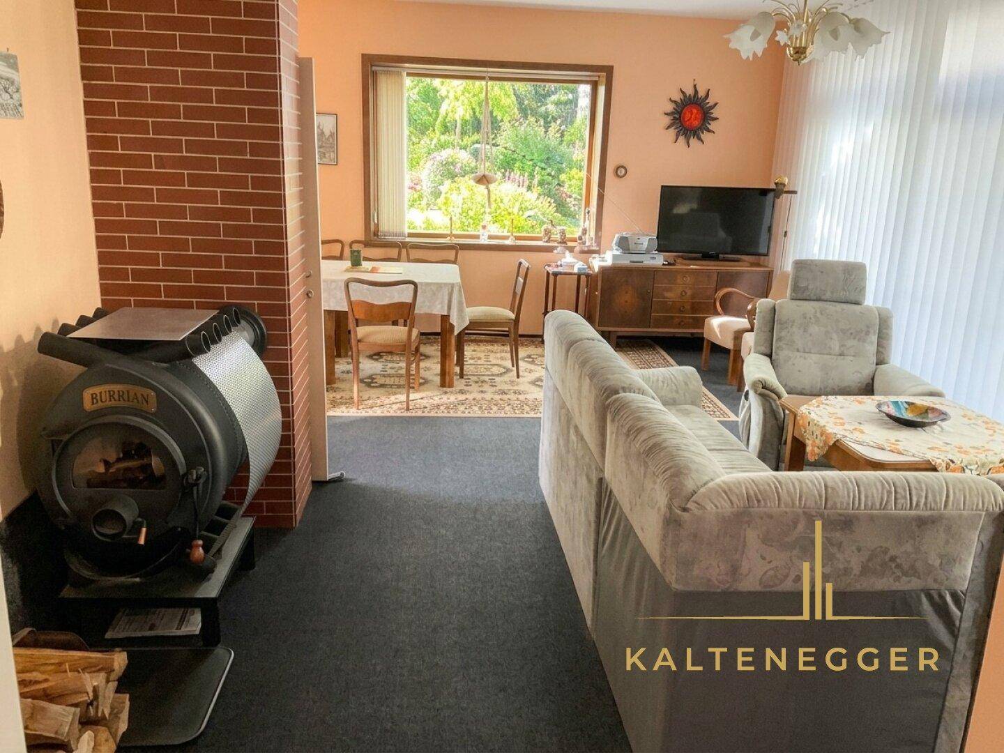 Wohn- und Essbereich mit einem Holzofen, gemütlichem Sofa und Blick ins Grüne durch das Fenster.
