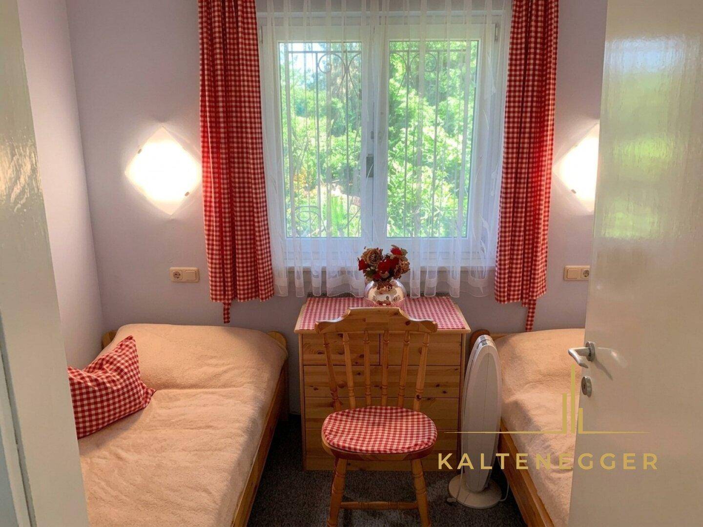 Kleines Schlafzimmer mit zwei Einzelbetten, Holzkommode und Fenster mit rot-weiß karierten Vorhängen.