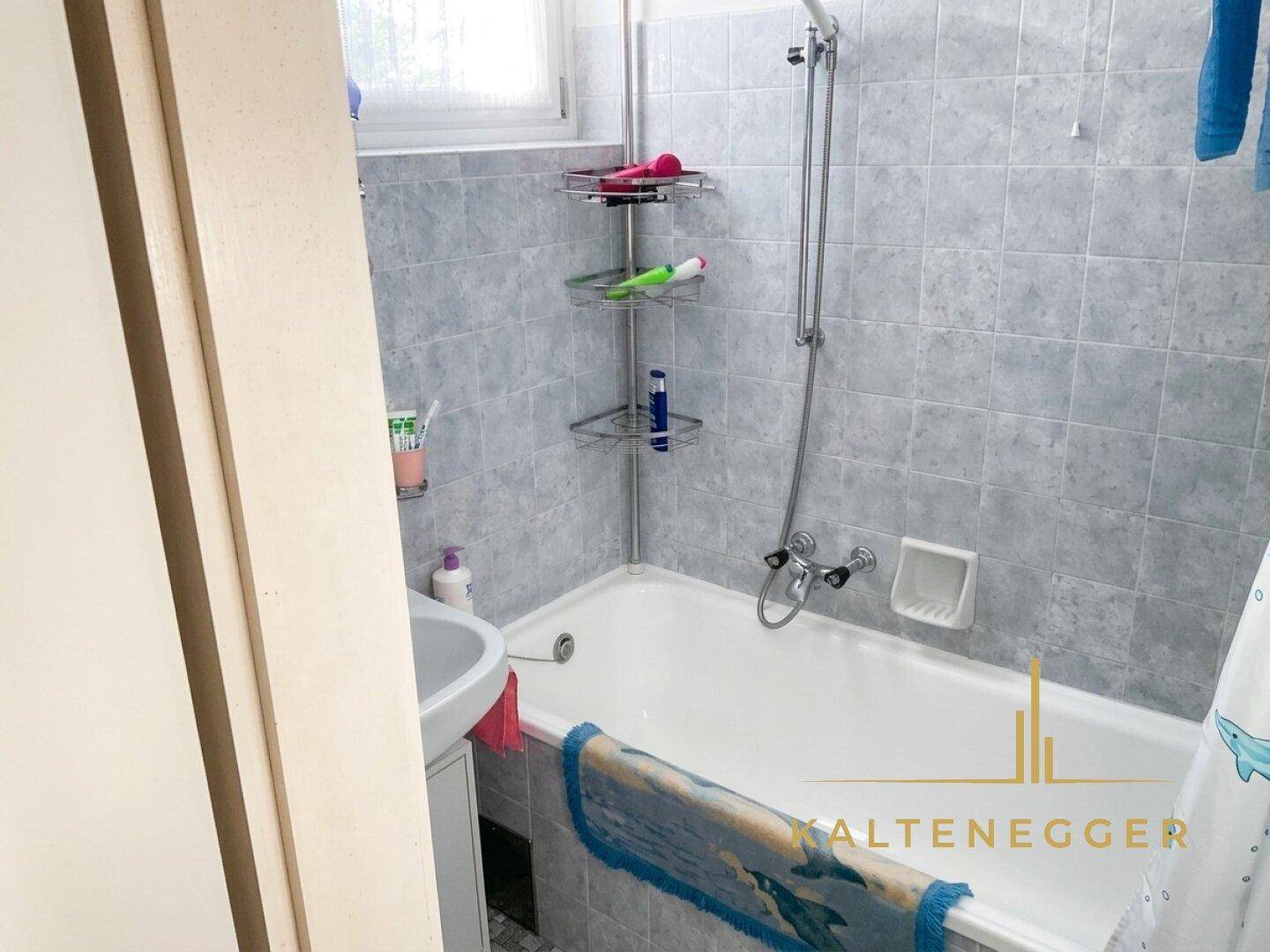 Badezimmer mit Badewanne, Waschbecken und grauen Fliesen, ausgestattet mit einem Fenster.