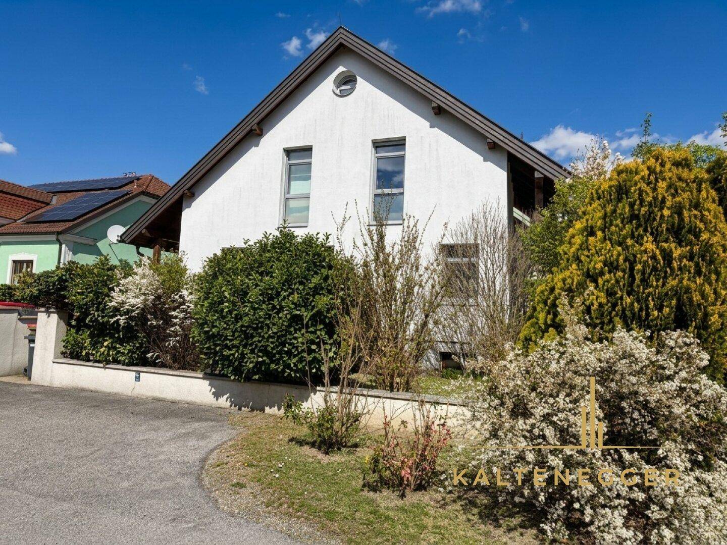 Seitenansicht des Hauses mit weißer Fassade, umgeben von blühenden Büschen und Sträuchern an einem sonnigen Tag.