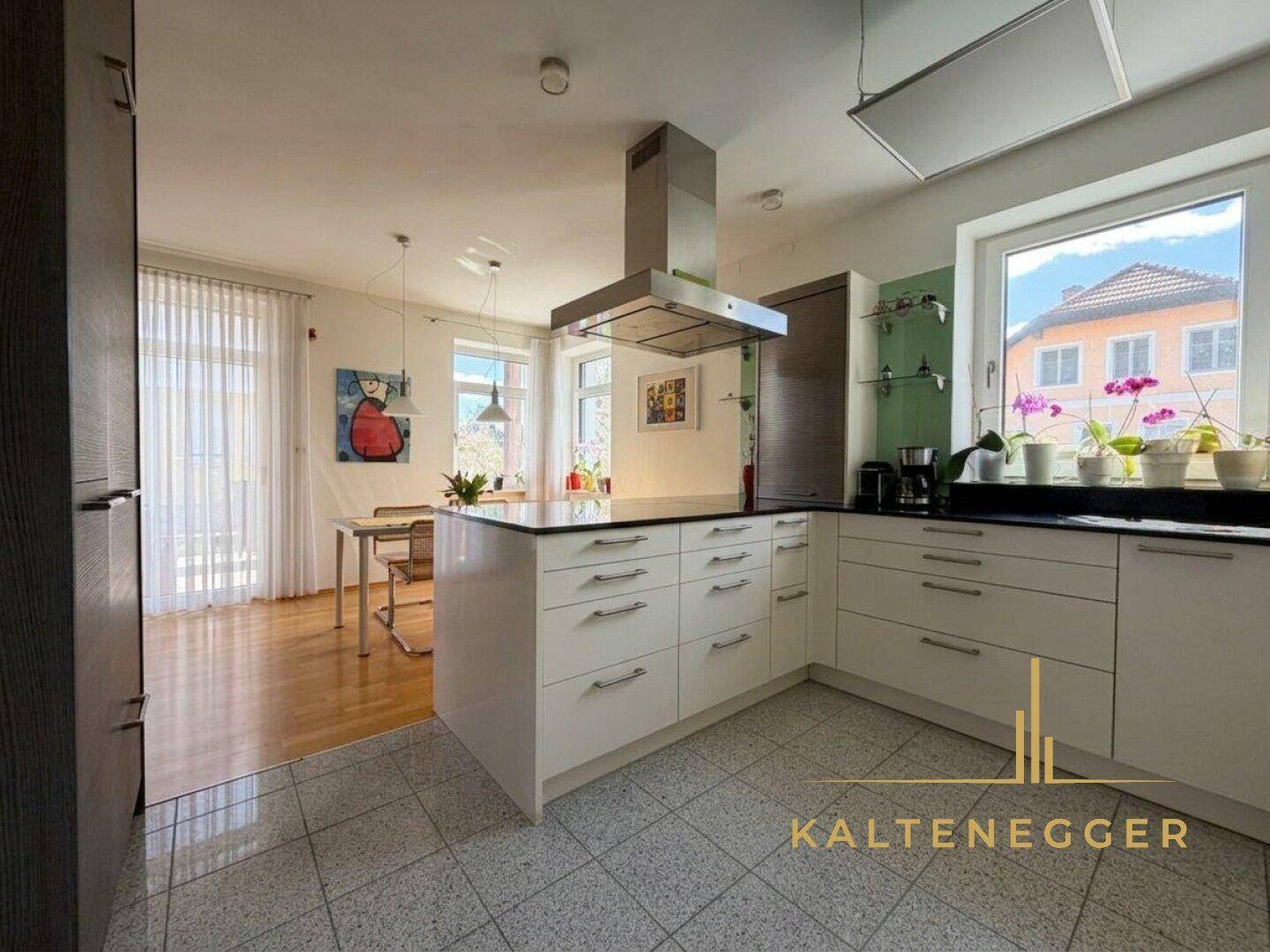 Moderne weiße Küche mit Kochinsel und Essbereich im Hintergrund, ausgestattet mit hochwertigen Geräten.