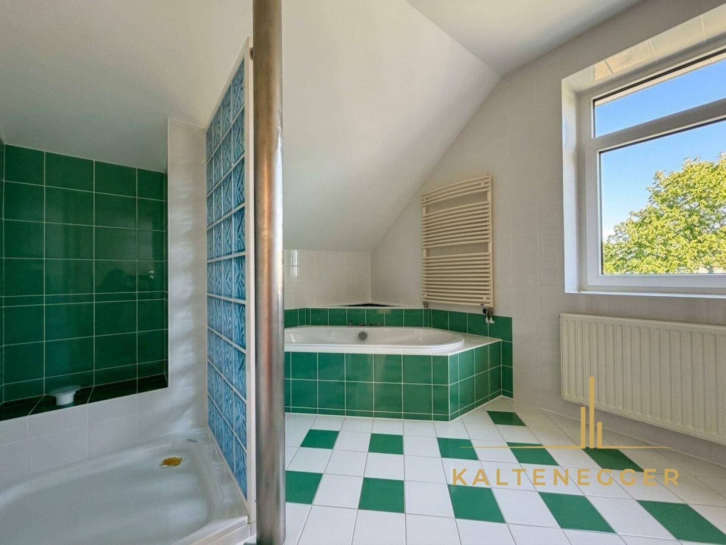 Badezimmer mit grünen und weißen Fliesen, Eckbadewanne, Dusche und Fenster unter Dachschräge.