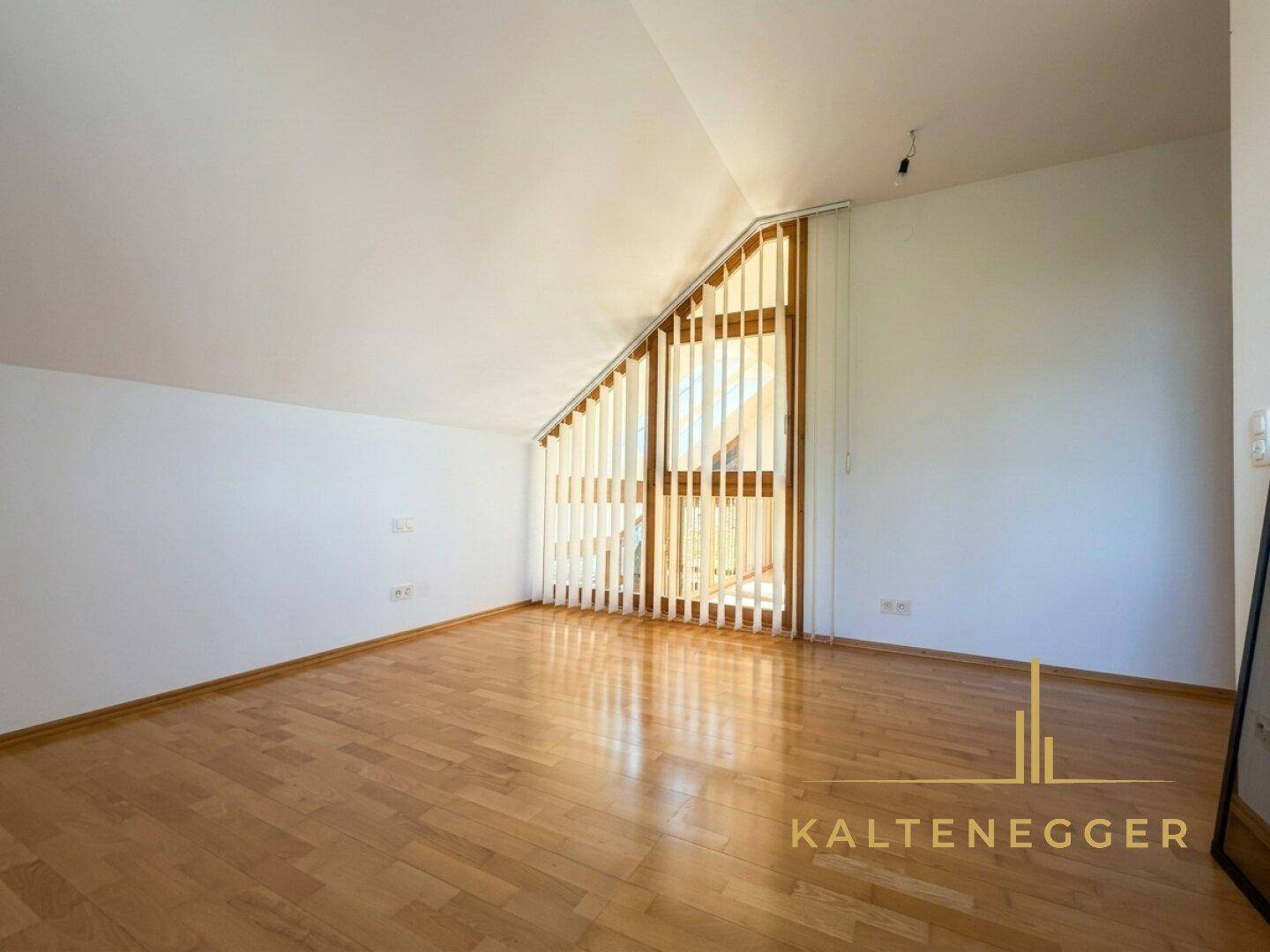 Heller Raum mit Holzboden und schrägen Wänden, ausgestattet mit einem großen Fenster und Jalousien.