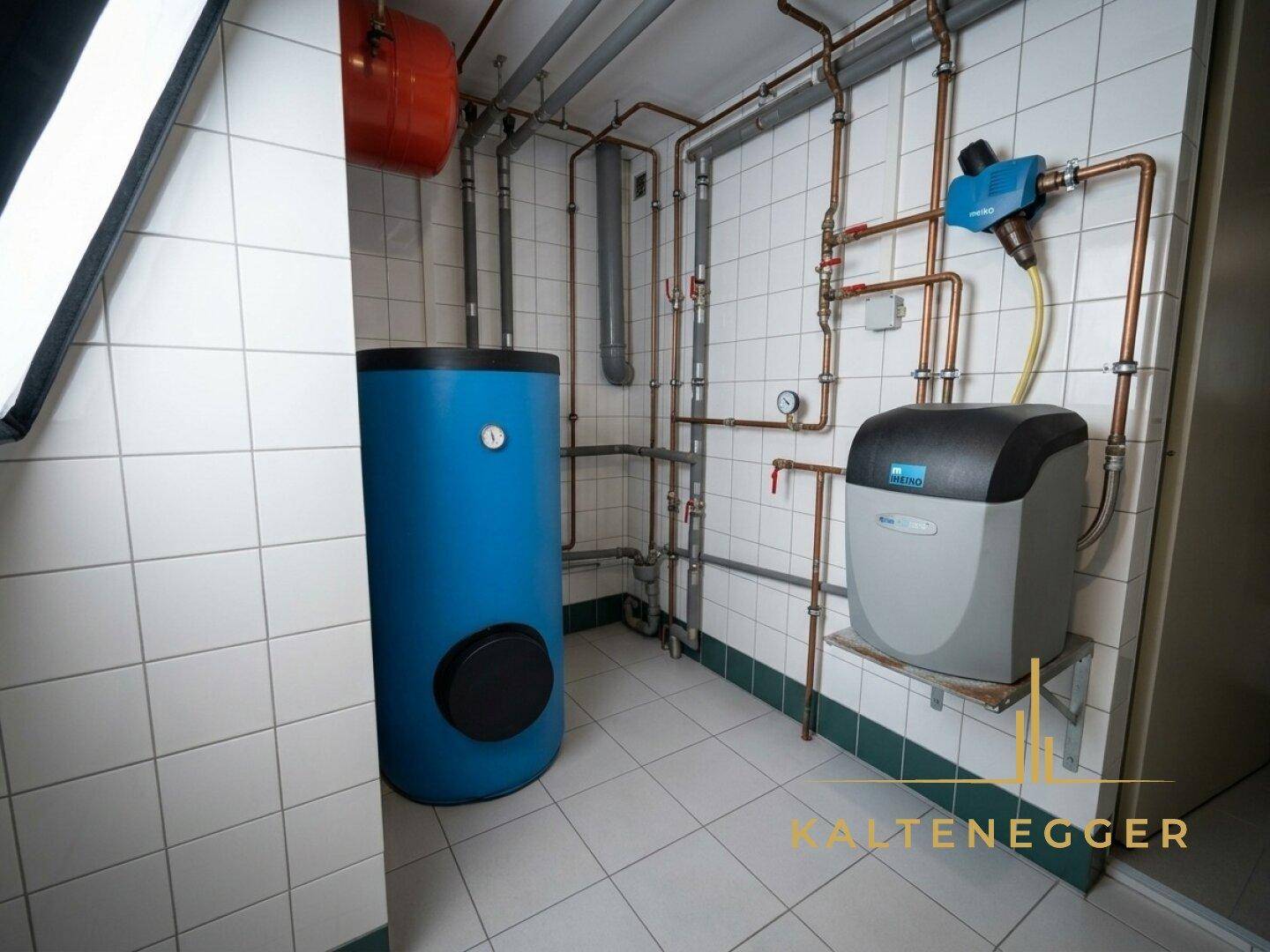 Technikraum mit Warmwasserspeicher, Wasserenthärtungsanlage und sichtbaren Rohrleitungen auf gefliestem Boden.