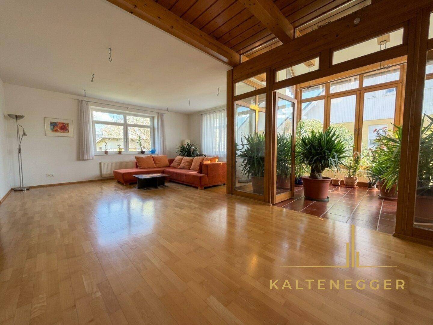 Helles Wohnzimmer mit Holzboden, sichtbaren Deckenbalken und einem lichtdurchfluteten Wintergartenbereich.