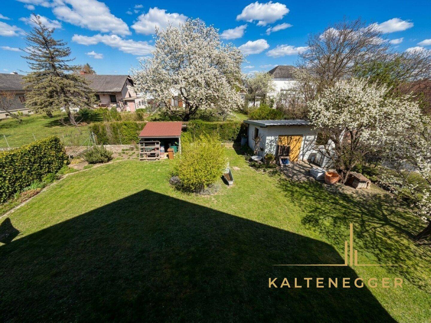 Weitläufiger Garten mit Rasenflächen, zwei Gartenhäusern und blühenden Bäumen im Frühling.