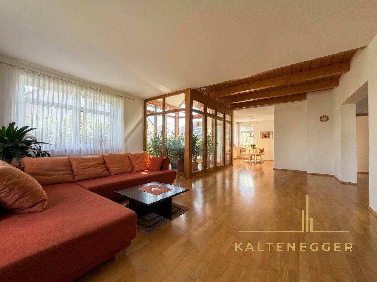 Geräumiges Wohnzimmer mit gemütlicher orangefarbener Couch, Holzbalkendecke und angrenzendem Wintergarten.
