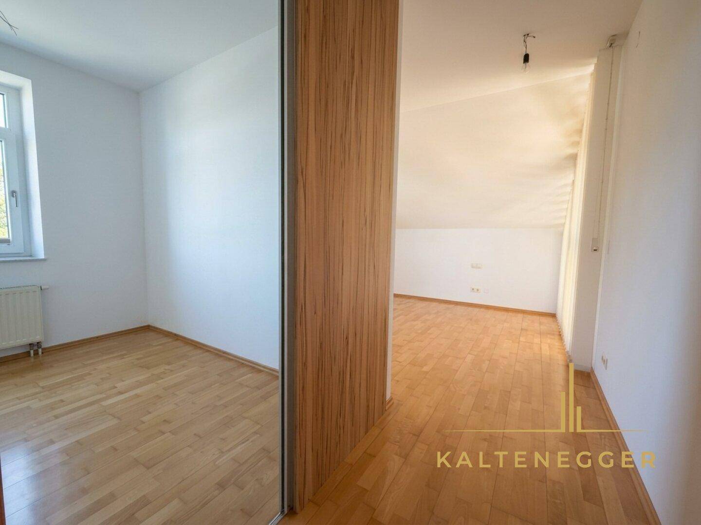 Zwei helle, leere Räume mit Holzboden und Dachschrägen, getrennt durch eine Holzwand mit Spiegel.