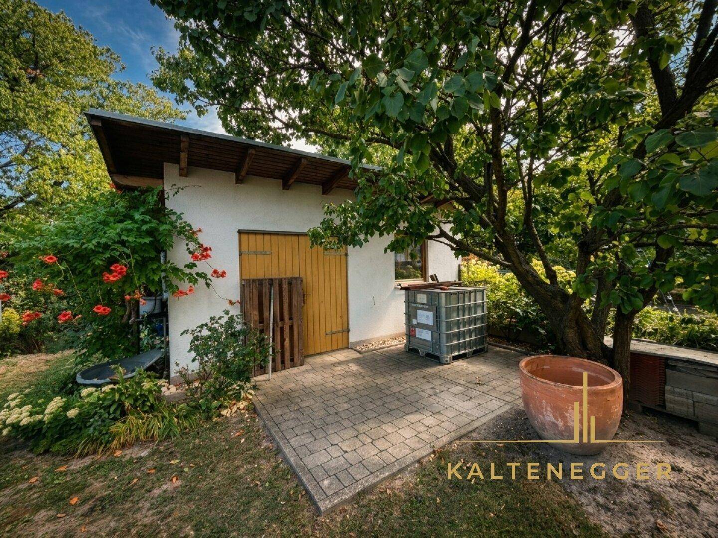 Kleines Gartenhaus mit gepflastertem Vorplatz, umgeben von Bäumen und üppigem Grün im Garten.