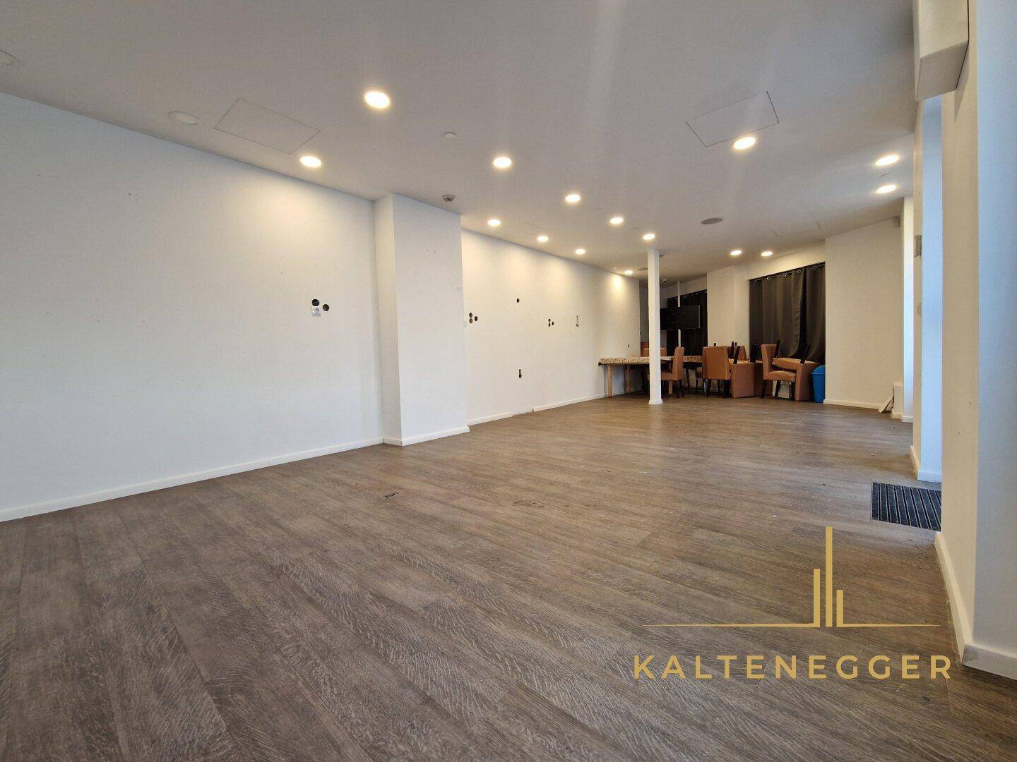 Großzügiger, leerer Verkaufsraum mit hellem Laminatboden, weißen Wänden und moderner Deckenbeleuchtung, bereit zur Nutzung.