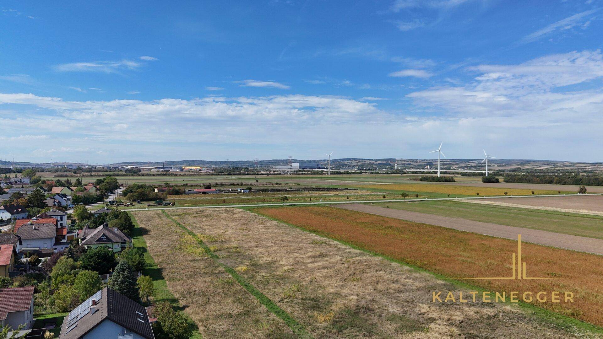 Panoramablick über weite Felder, ländliche Umgebung und Windräder am Horizont unter blauem Himmel.