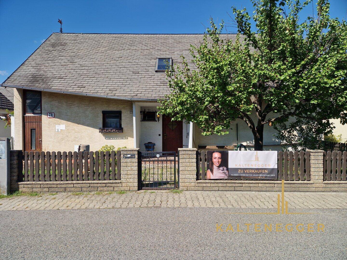 Vorderansicht des Hauses von der Straße mit Zaun und einem "Zu Verkaufen"-Schild.