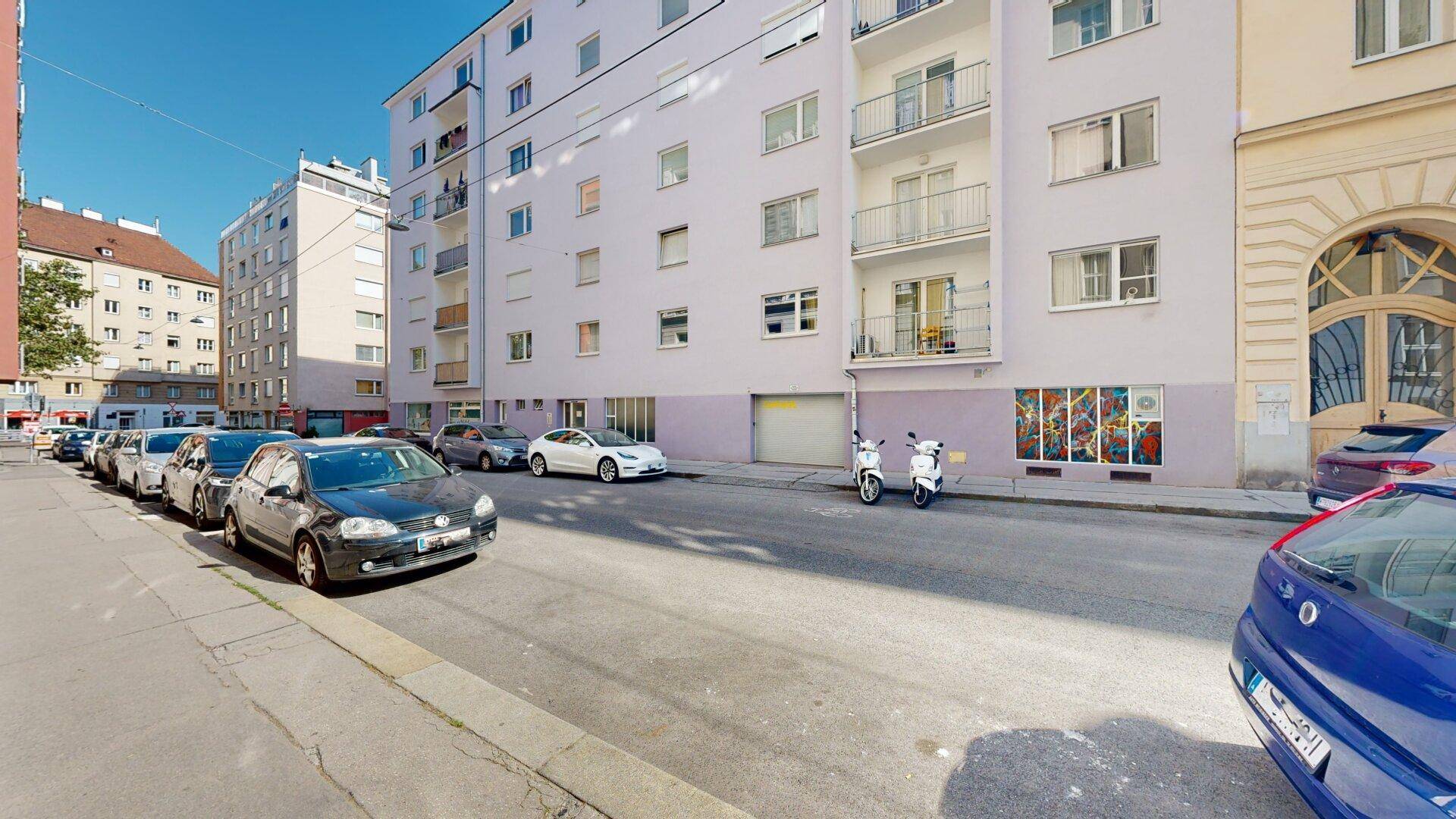 Seitenansicht eines Wohngebäudes mit Balkonen und geparkten Autos auf der Straße.