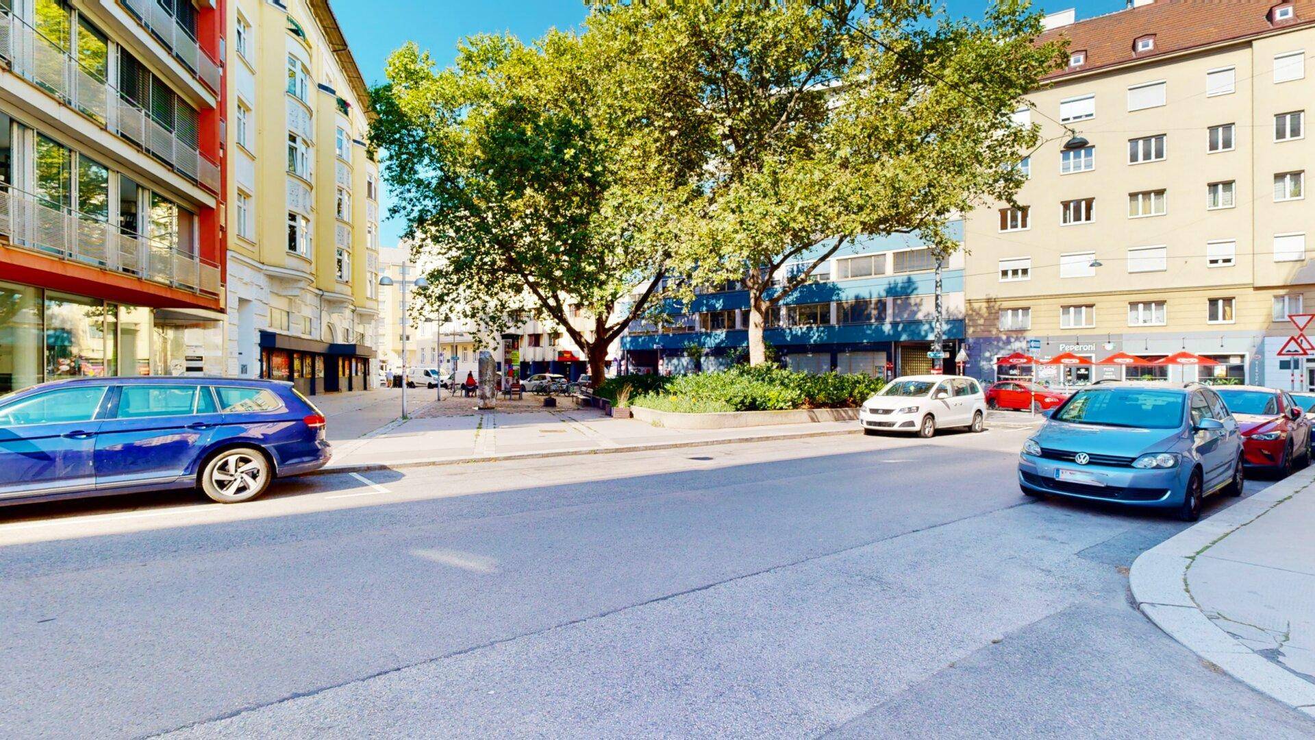 Belebtes Stadtbild mit Bäumen, parkenden Autos und Gebäuden im Hintergrund.