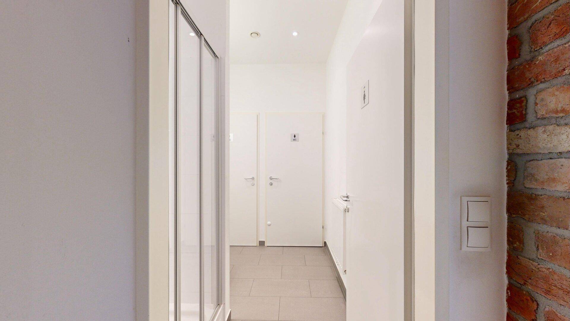 Heller Flur mit Fliesenboden und einer Duschkabine, der zu den Toiletten führt.