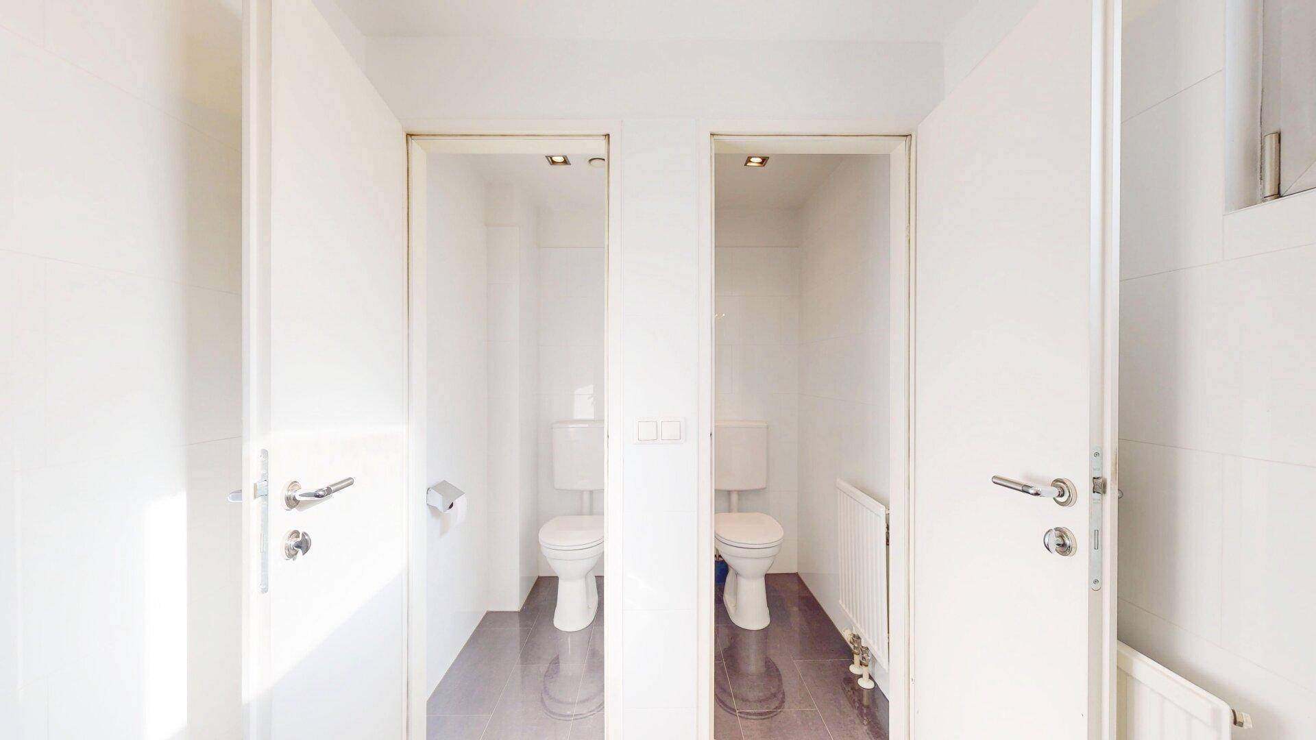 Zwei separate Toilettenkabinen mit weißen Toiletten und grauen Fliesen, sauber und funktional.