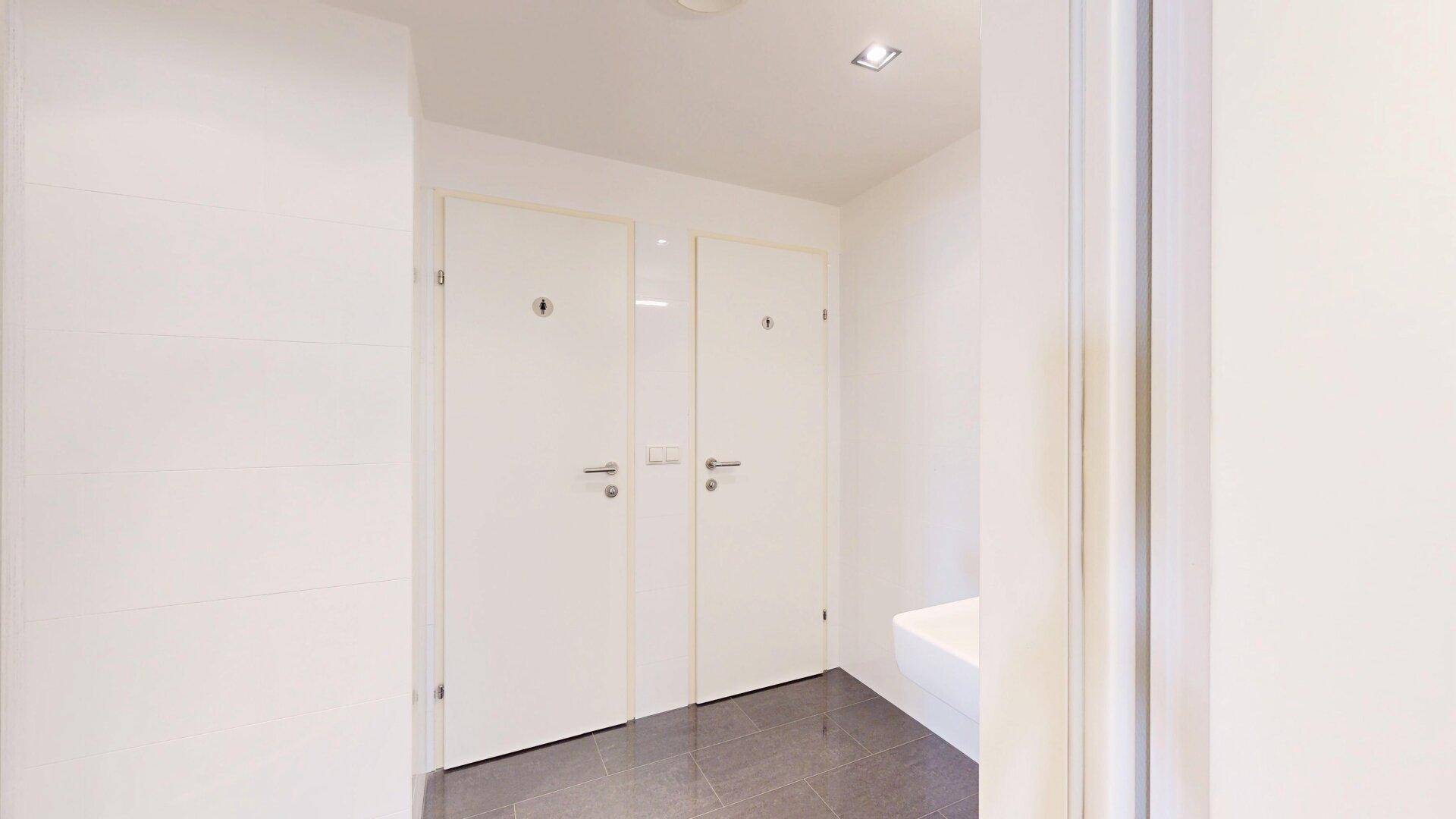Heller Flur mit grauen Fliesen, der zu den Toiletten führt, mit weißen Türen.