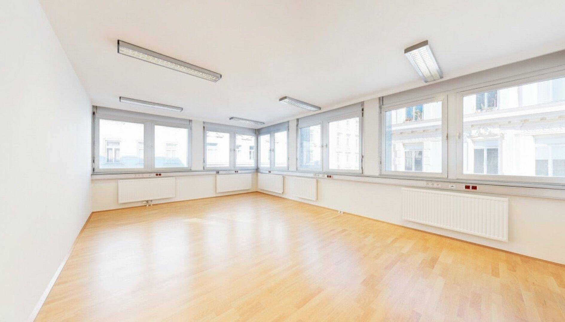 Geräumiges, leeres Büro mit großen Fenstern, Heizkörpern und Holzfußboden, ideal für flexible Nutzung.
