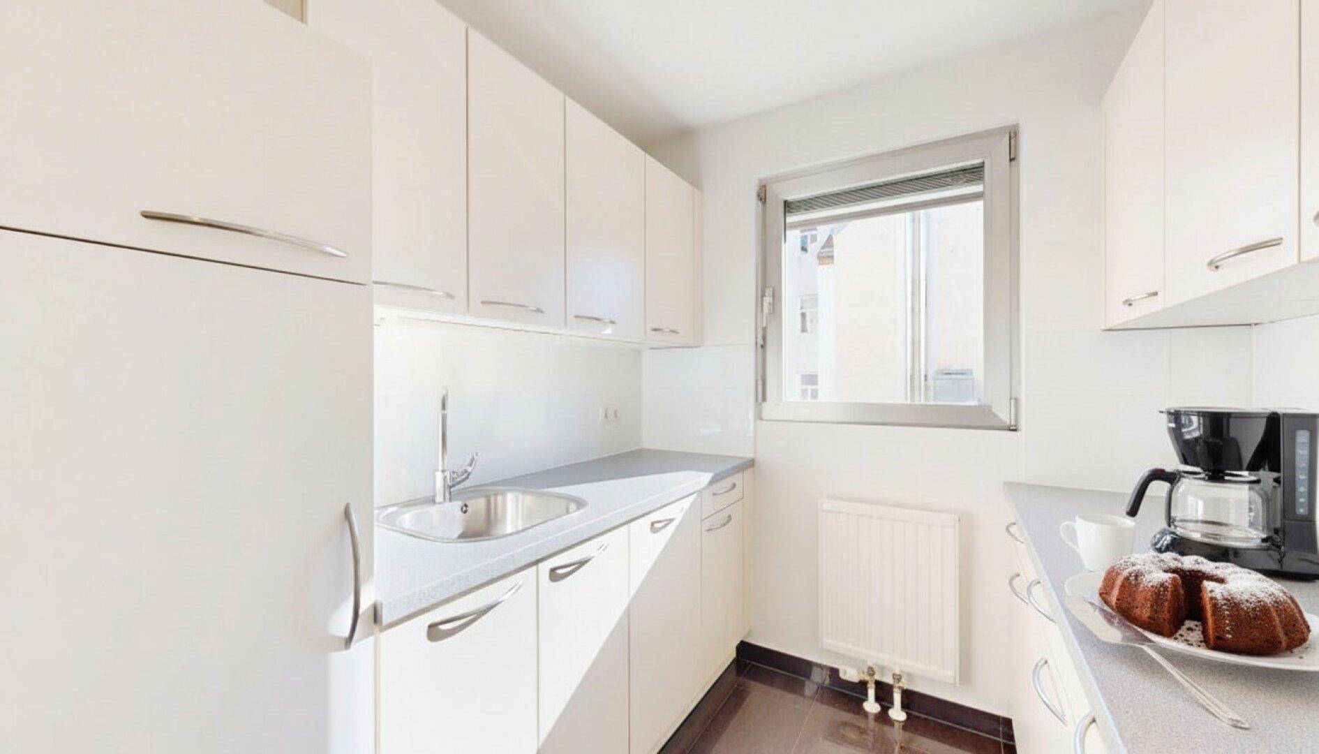 Funktionale Küchenzeile mit weißen Schränken, Spüle, Kühlschrank und Kaffeemaschine, mit Fenster.