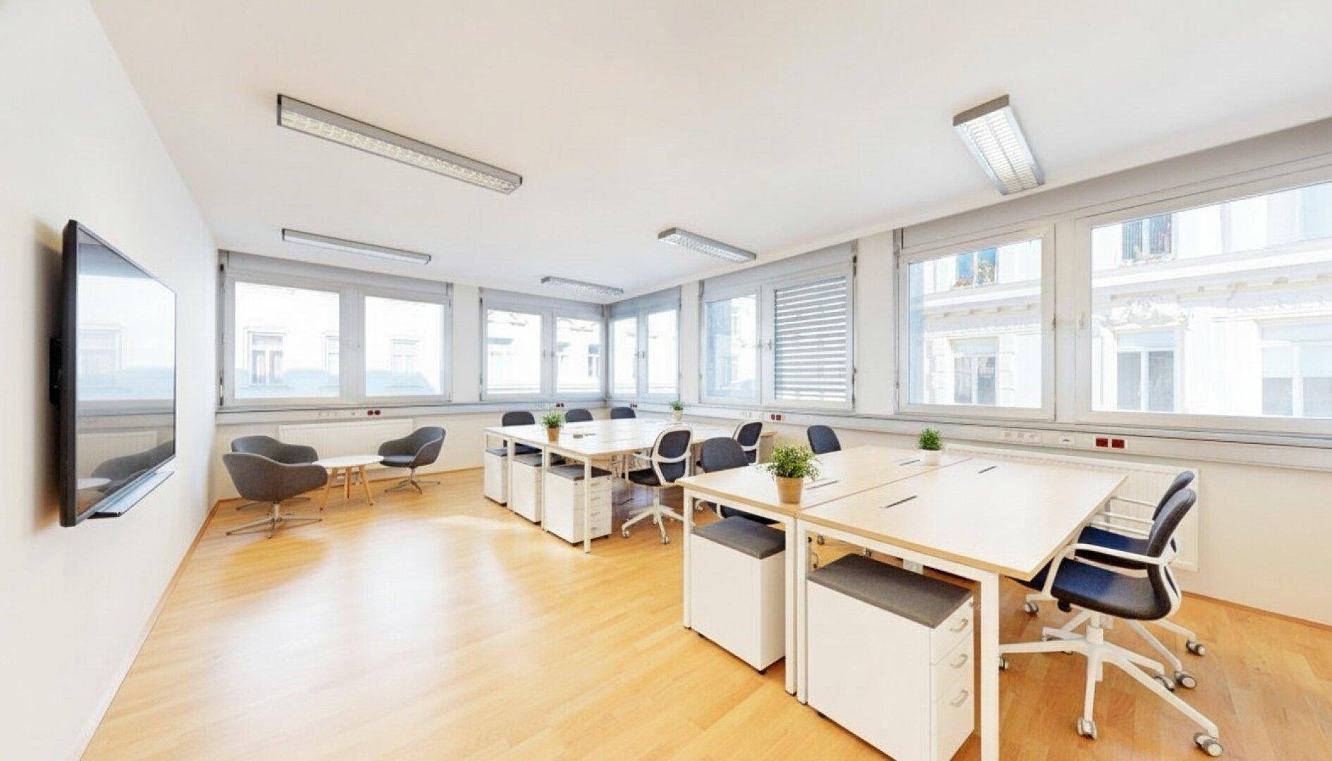 Modern eingerichtetes Büro mit mehreren Arbeitsplätzen, Wandfernseher und großen Fenstern mit Jalousien.