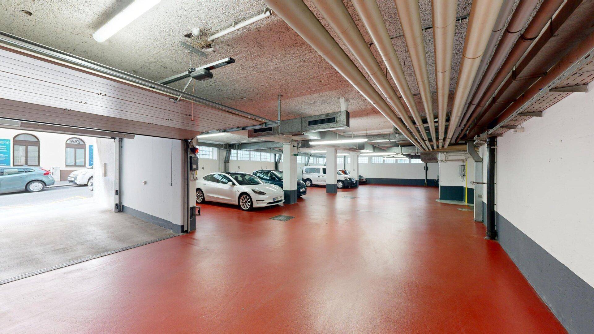 Geräumige Tiefgarage mit rotem Boden und sichtbaren Versorgungsleitungen an der Decke, mehrere Autos parken.