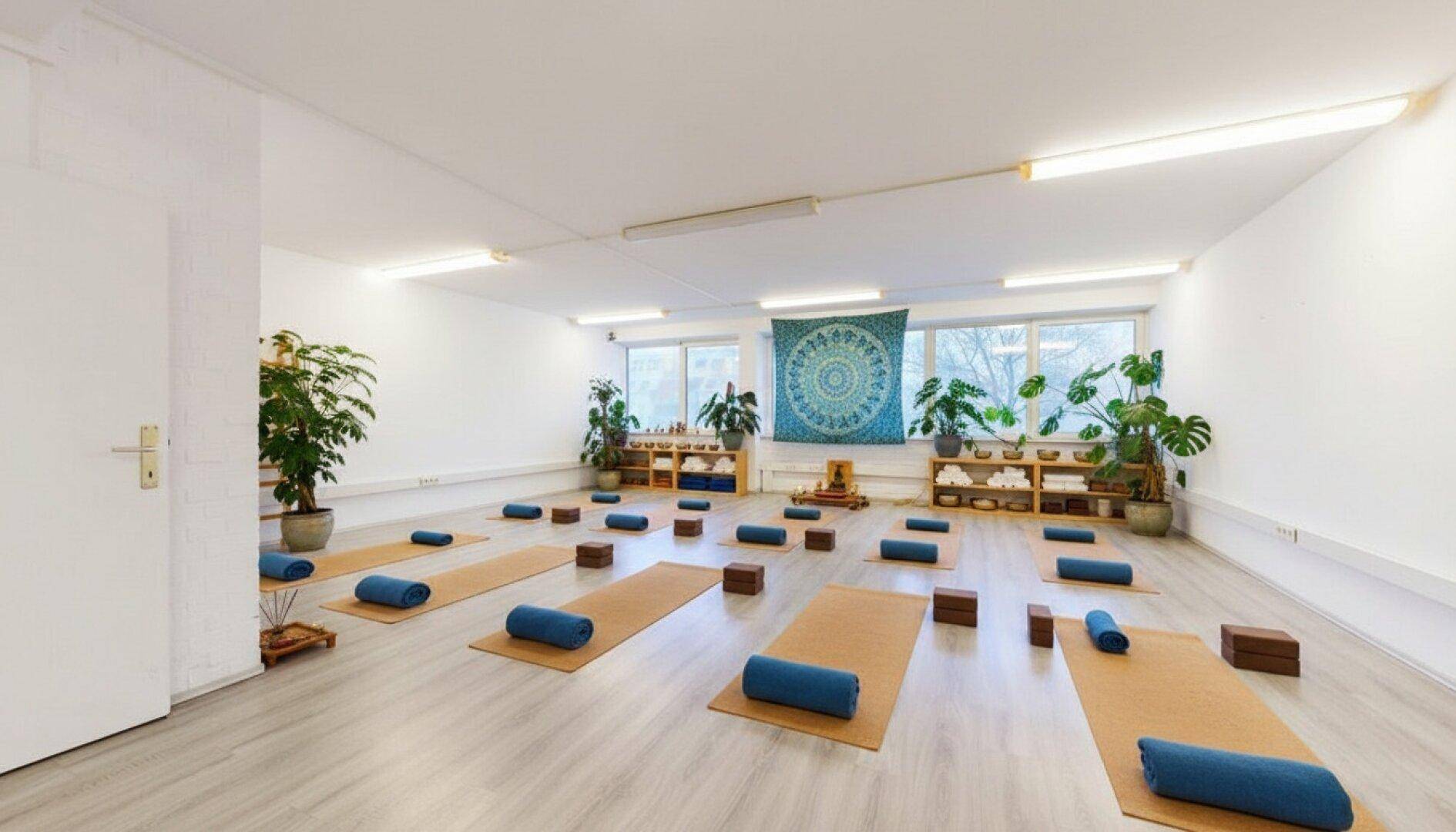 Ruhiger Yogaraum mit Matten, Pflanzen und einem Wandteppich, ideal für Entspannung und Kurse.