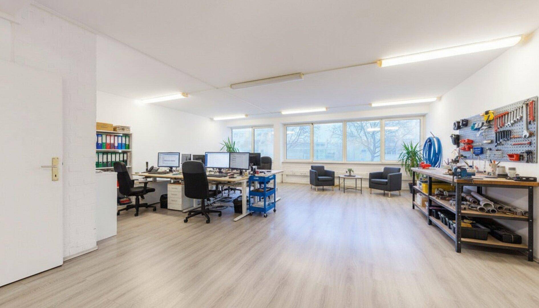 Modern eingerichteter Büroraum mit mehreren Arbeitsplätzen, einer Sitzecke und einem Werkstattbereich, hell und funktional.