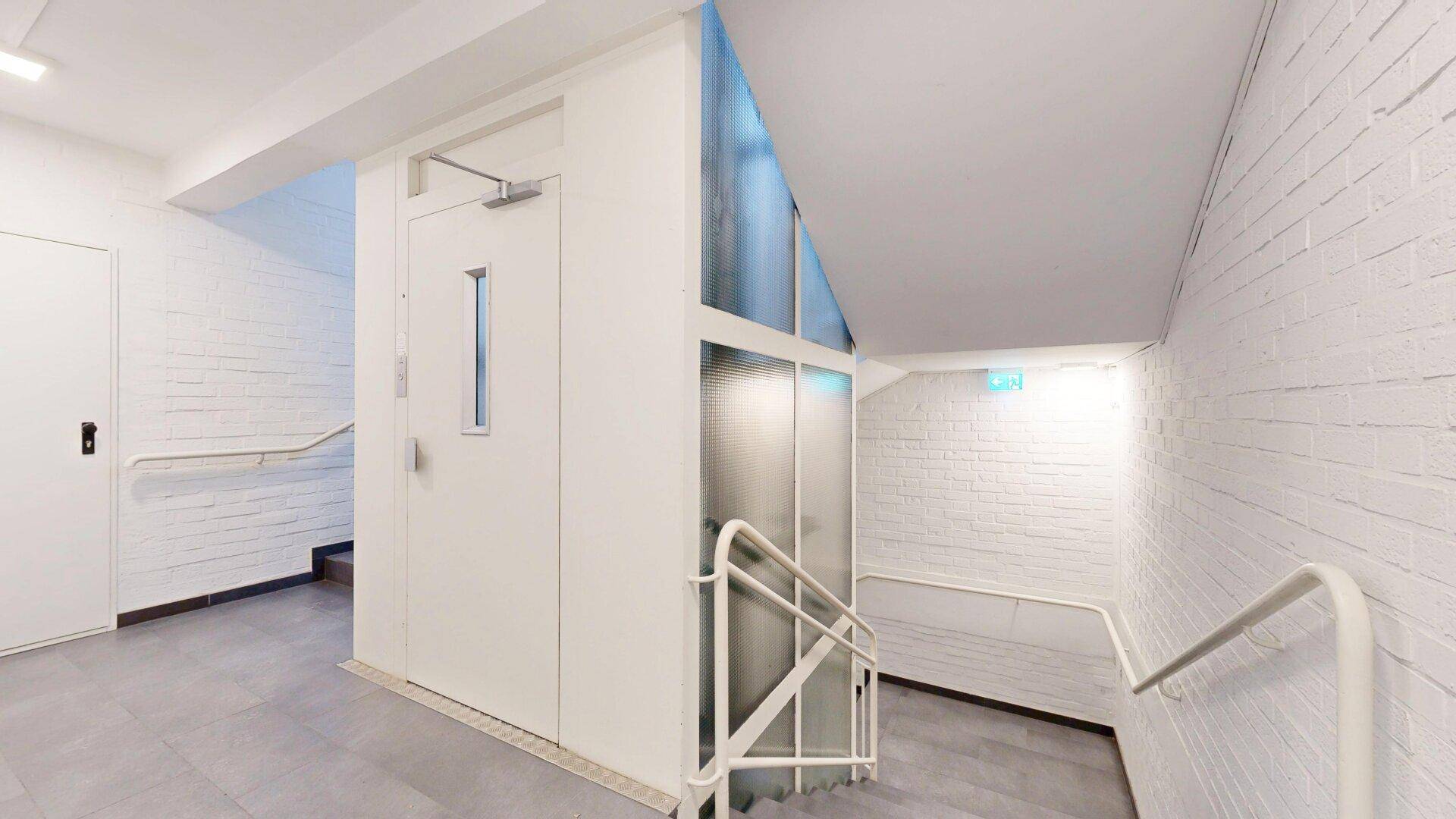 Treppenhaus mit Aufzug und Handläufen, weißen Ziegelwänden und grauem Fliesenboden, gut beleuchtet.