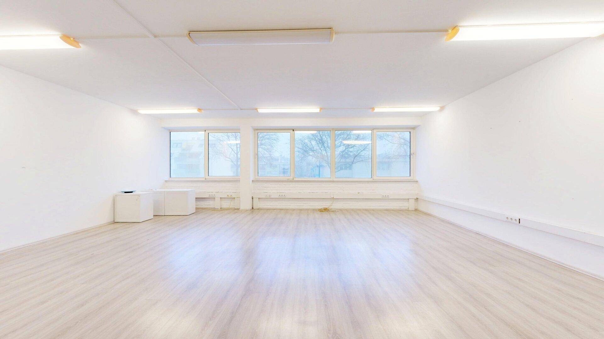 Weitläufiger, heller Büroraum mit großen Fenstern, die viel Tageslicht hereinlassen, und einem modernen Laminatboden.