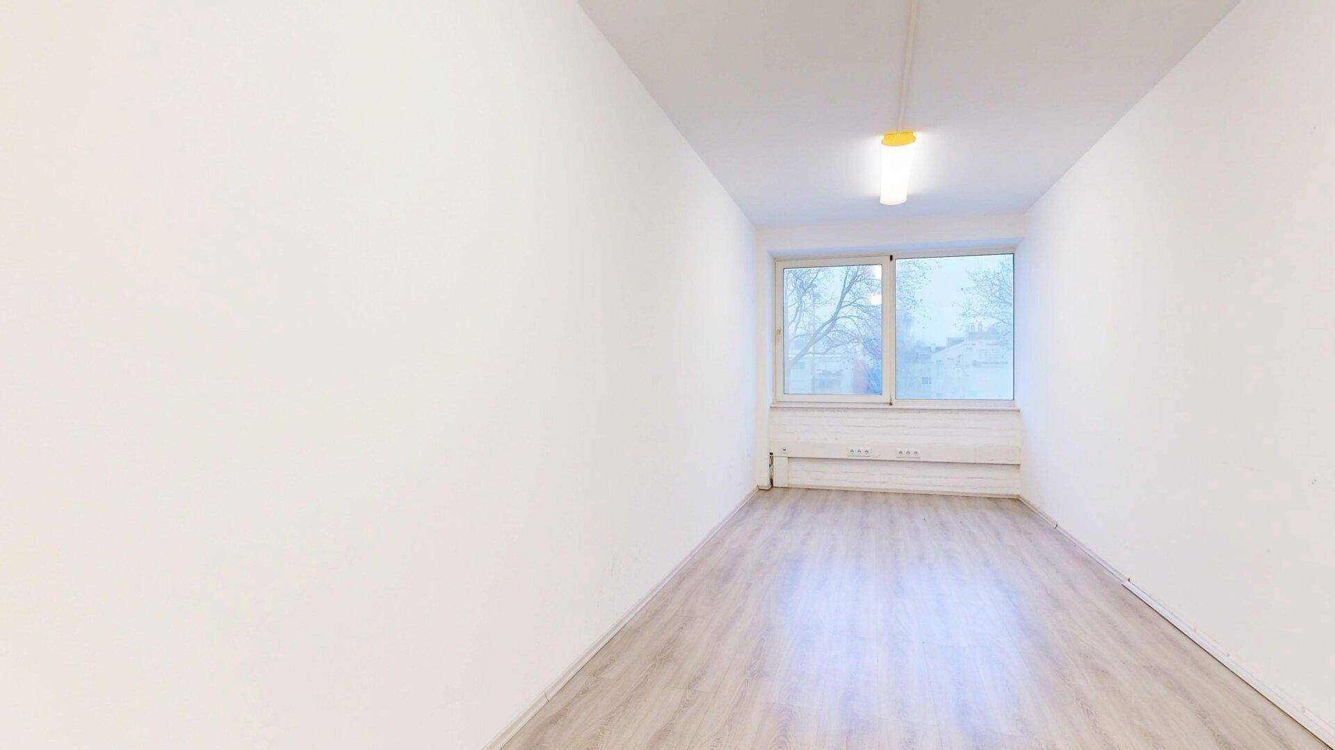 Schmaler, heller Büroraum mit weiß gestrichenen Wänden, Laminatboden und einem Fenster am Ende des Raumes.