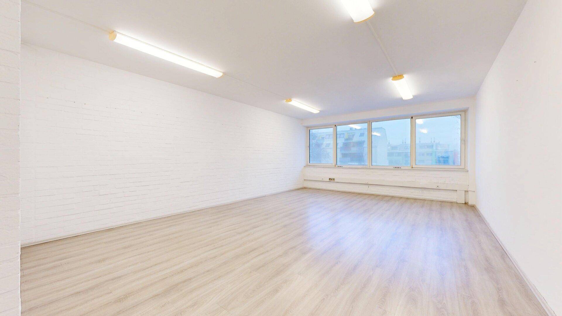 Heller, leerer Büroraum mit einer weißen Ziegelwand, Laminatboden und großen Fenstern, die viel Licht spenden.