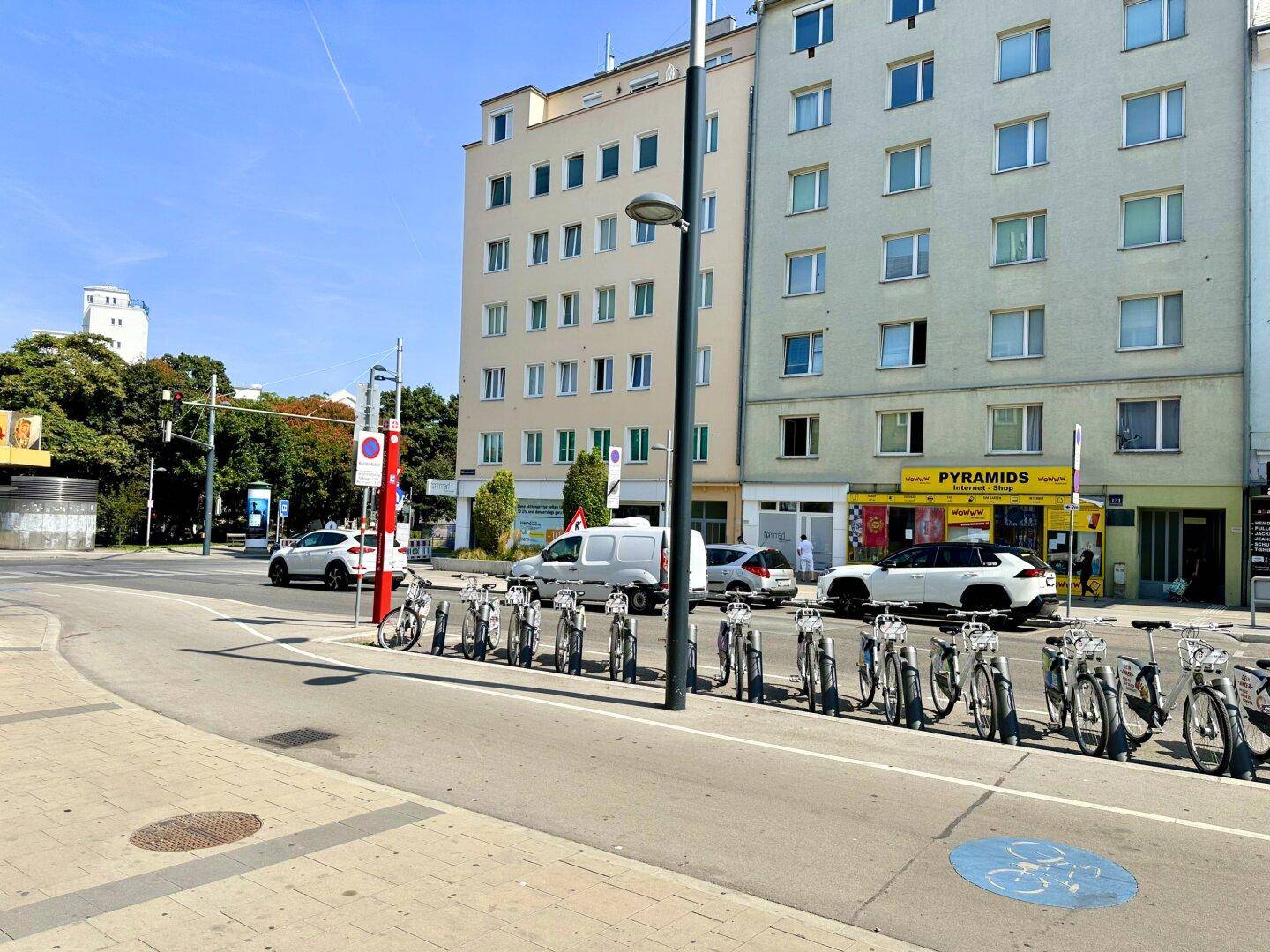 Straßenansicht mit modernen Gebäuden, parkenden Autos und Fahrradständern unter blauem Himmel.
