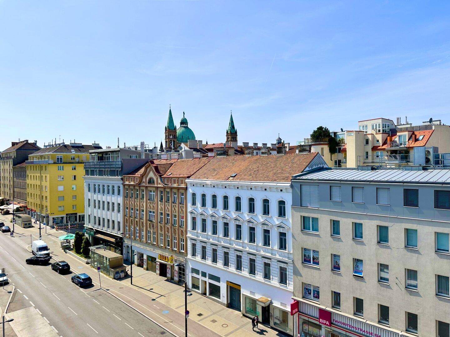 Panoramablick über eine belebte Straße mit verschiedenen Gebäudefassaden und einem Kirchturm im Hintergrund.