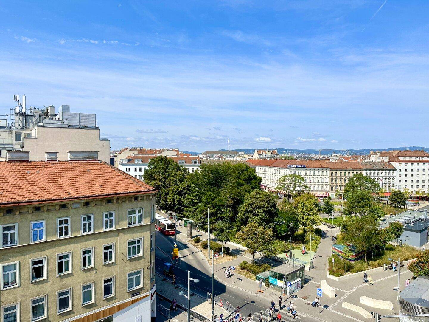 Weitläufiger Blick über eine Stadtkreuzung mit viel Grün und Gebäuden im Hintergrund.