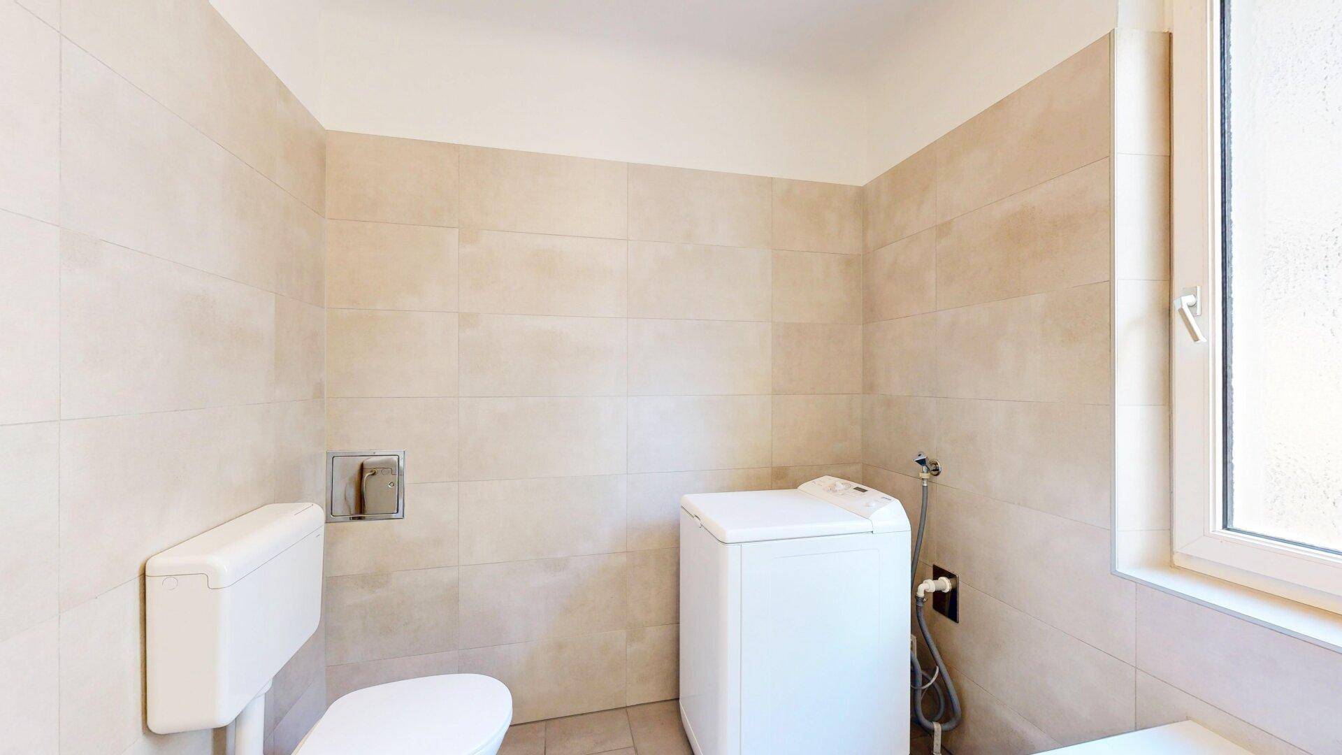 Badezimmerbereich mit WC, Waschmaschine und hellem Fliesenboden.