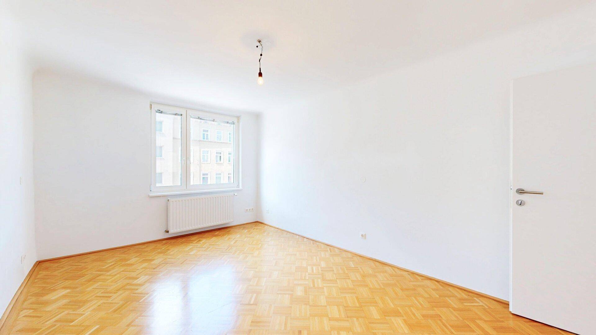 Großes Schlafzimmer mit Fischgrätparkett und einem Fenster, das den Raum hell erscheinen lässt.