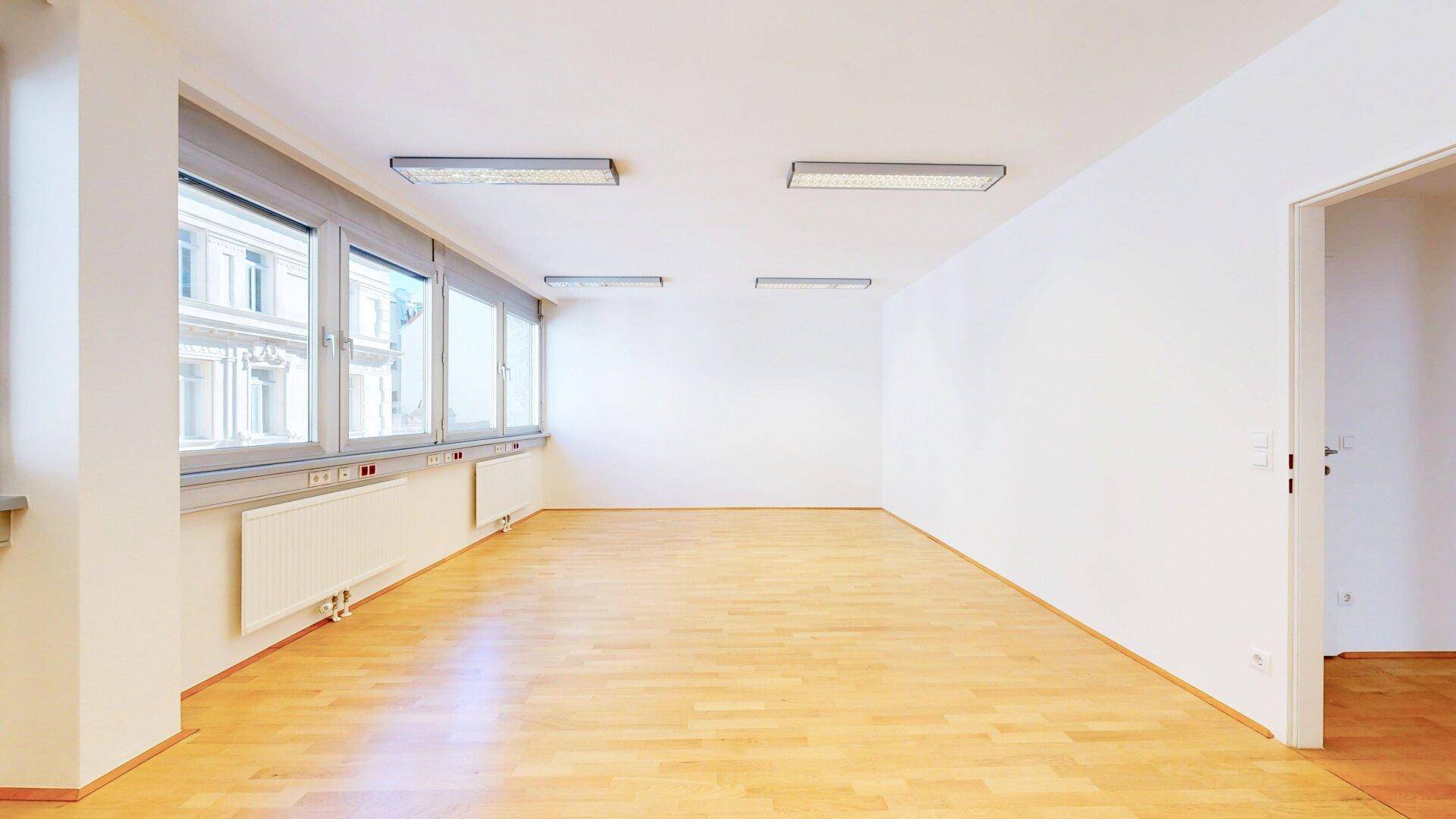Helles, leeres Büro mit großen Fenstern, Heizkörpern und Holzfußboden, bereit zur Einrichtung.