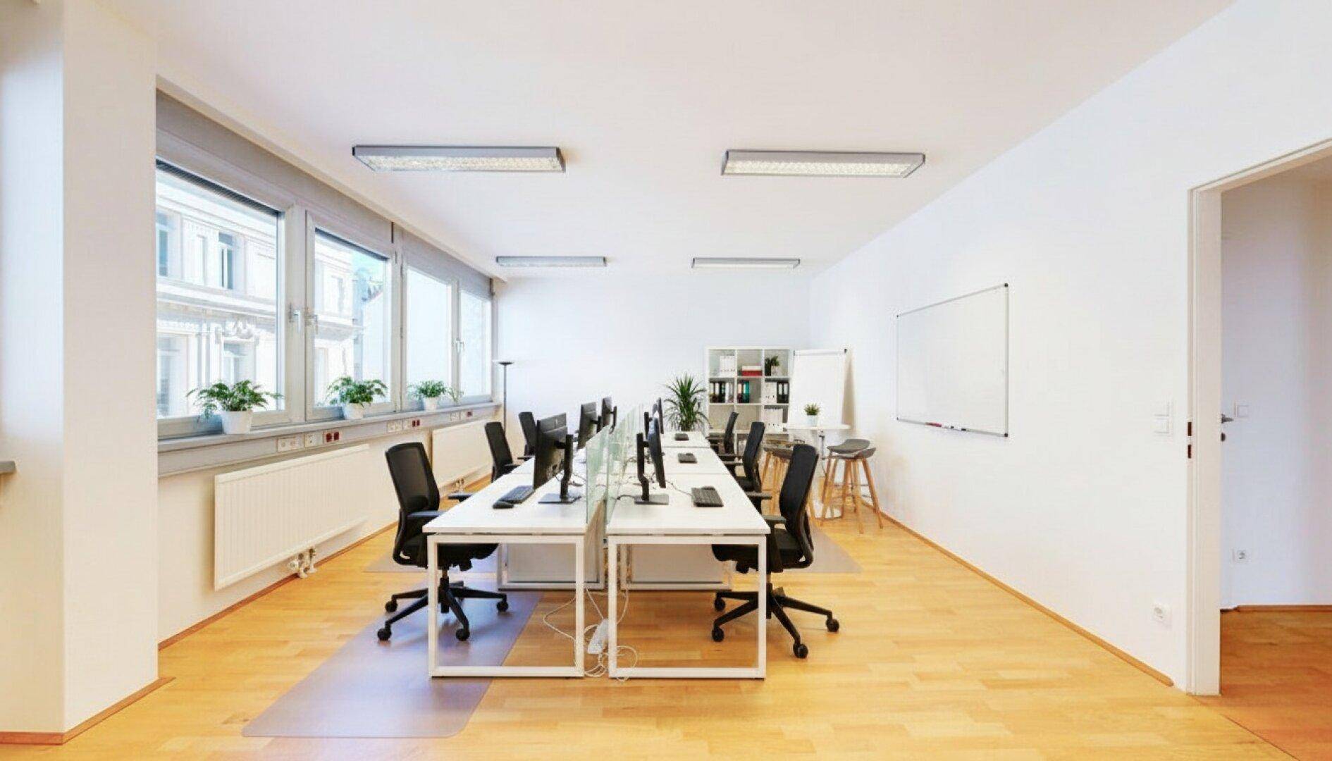 Modern eingerichtetes Großraumbüro mit mehreren Arbeitsplätzen, Pflanzen und großen Fenstern.