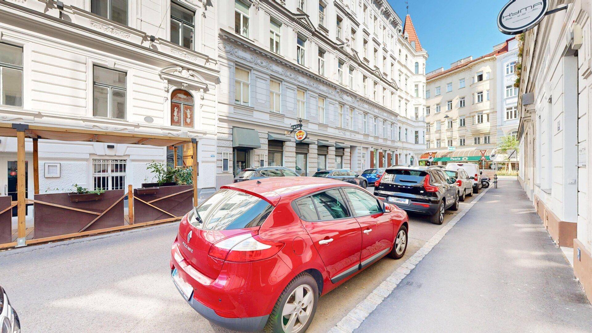 Belebte Stadtstraße mit geparkten Autos und historischen Gebäudefassaden unter blauem Himmel.