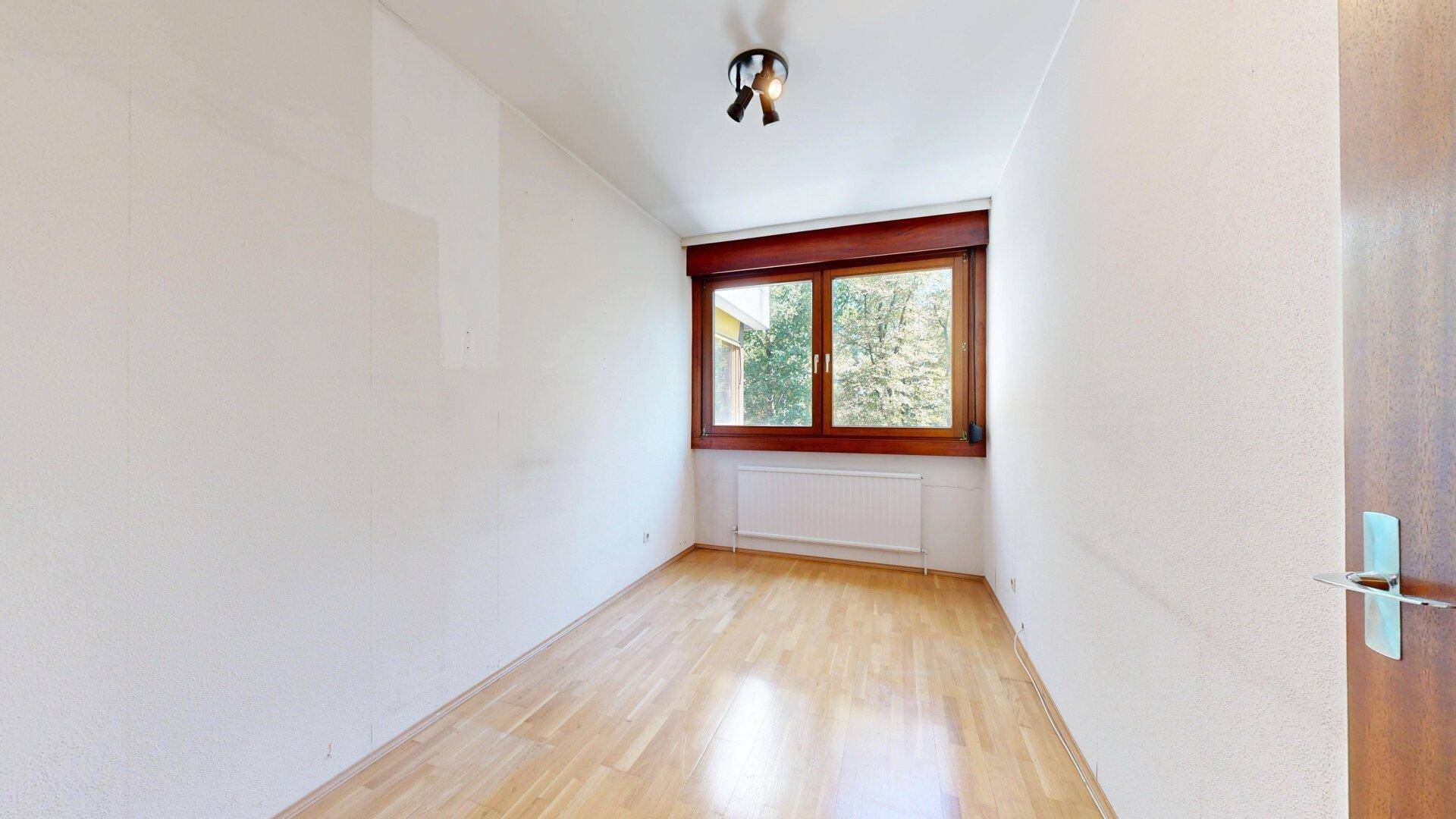 Leeres Schlafzimmer mit hellem Parkettboden und Fenster zum Außenbereich.