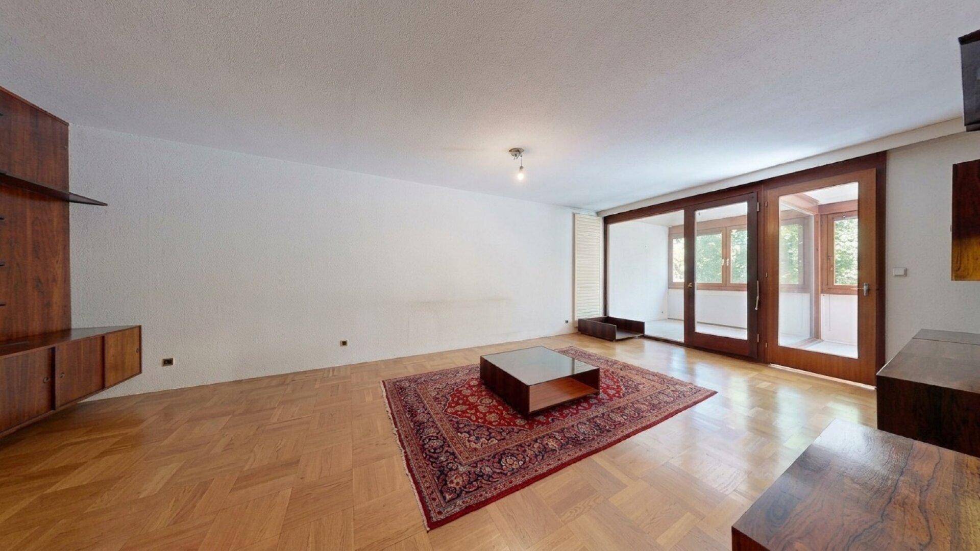 Geräumiges Wohnzimmer mit Fischgrätparkett, Teppich und großen Fenstern zum Außenbereich.