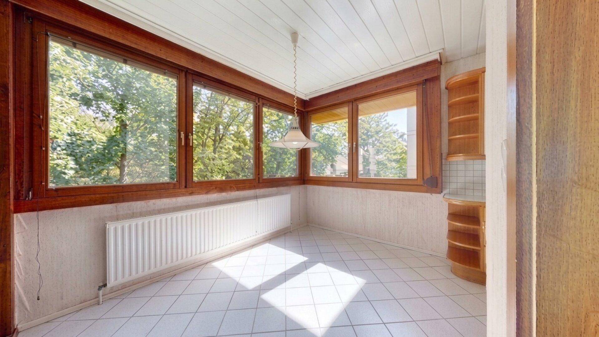 Sonniger Wintergarten mit großen Fenstern, Heizkörper und hellem Fliesenboden.