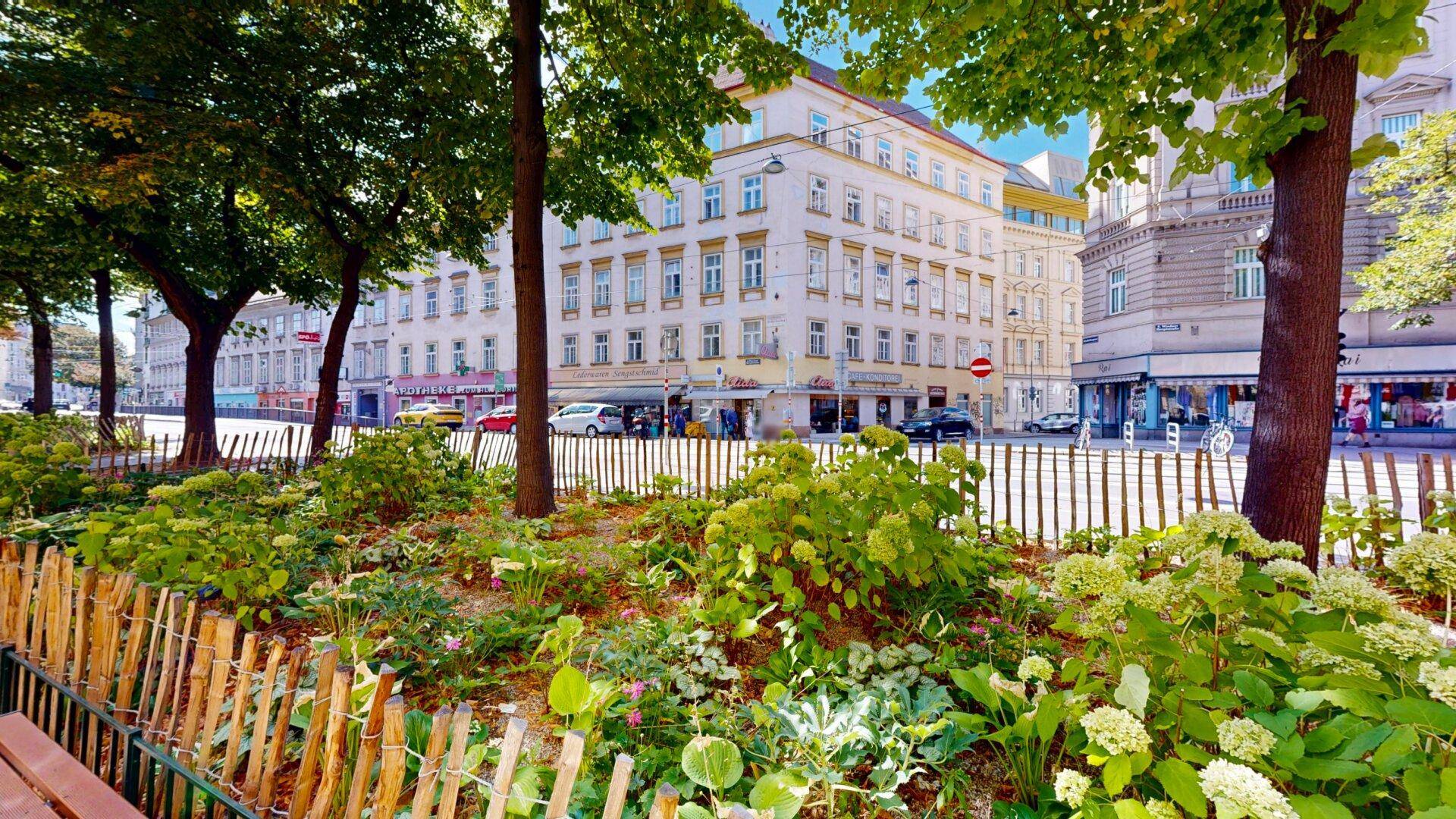 Grüne Parkanlage mit Bäumen und Blumenbeeten vor einer belebten Stadtstraße mit Gebäuden.