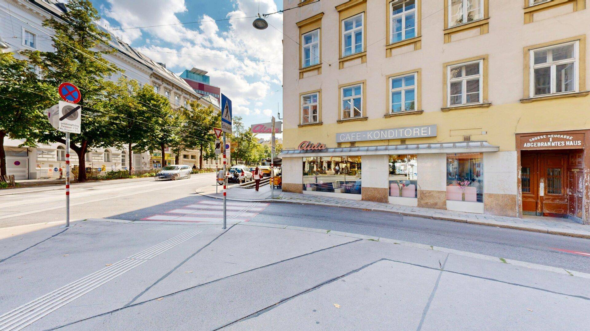 Straßenecke mit einem Café, historischen Gebäuden und Bäumen unter blauem Himmel.